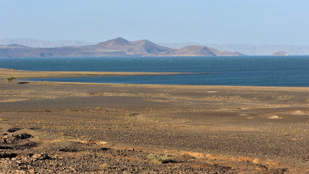 Arid, rocky foreground leading to the blue waters of Lake Turkana, with volcanic hills and mountains in the distance under a clear sky.